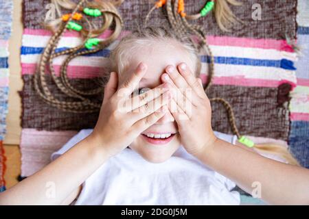 Primo piano ritratto di ragazza allegra con i pigtail giacenti sul pavimento su un tappeto intrecciato a righe, sorridente, giocando a nascondino e cerca. Foto Stock