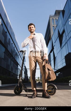 vista ad angolo basso di un uomo d'affari con cuscinetti in regolazione formale dell'usura allacciare tenendo la borsa in pelle e stando in piedi vicino allo scooter elettrico Foto Stock