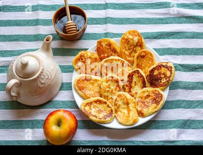 Composizione piatta con gustose frittelle fatte in casa disposte sul piatto vicino ad una mela, una teiera e una ciotola di miele. Focalizzazione selettiva sui pancake Foto Stock