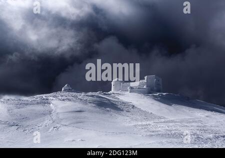 Il vecchio osservatorio coperto di neve testurizzata. Cielo drammatico e tempestoso con nuvole grigie. Scenario invernale. Splendido paesaggio di alte montagne. Sfondo Foto Stock