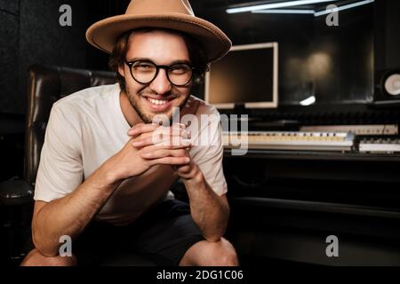 Attraente ragazzo sorridente gioiosamente posingon fotocamera in studio di registrazione del suono. Giovane musicista di stile che lavora felicemente in studio Foto Stock