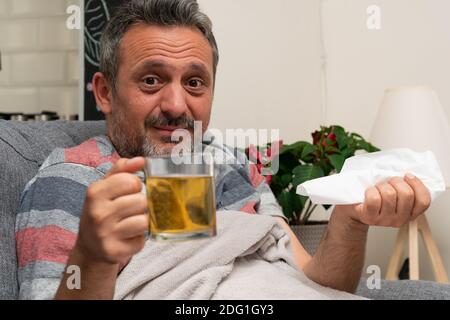 Uomo adulto con espressione preoccupata che si sente malbene avere influenza fredda sintomi di infezione da virus influenzale che presentano vetro trasparente tè caldo mag e pisolino Foto Stock