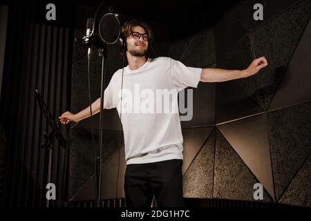 Giovane e attraente cantante maschile in cuffia che danzano durante la registrazione di una nuova canzone in studio moderno. Giovane musicista che canta in microfono Foto Stock