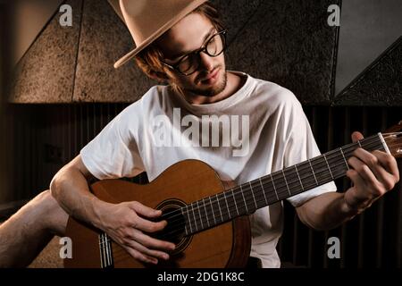 Ritratto di un attraente musicista maschile che gioca intentemente alla chitarra in studio di registrazione. Bell'elegante ragazzo che compone la canzone in un moderno studio audio Foto Stock