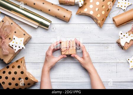 Scatole regalo di Natale e carta da imballaggio in colori oro su sfondo di legno bianco. Copiare spazio per il testo. Disposizione piatta e vista dall'alto. Mani femminili Foto Stock