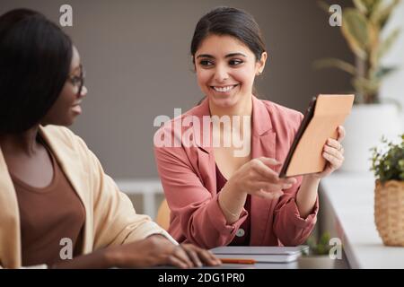 Ritratto di sorridente donna d'affari mediorientale guardando la collega femminile e puntando al tablet, mentre la gestione di affari di successo insieme, copia Foto Stock