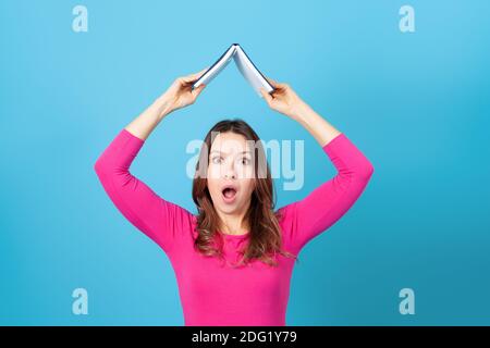 mock in su, una giovane donna sorpresa con la sua bocca aperta tenendo un diario o libro sopra la sua testa come un tetto, isolato su uno sfondo blu Foto Stock