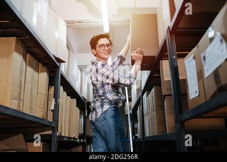 Felice lavoratore di magazzino femminile mettendo scatola di cartone pesante su un ripiano superiore Foto Stock