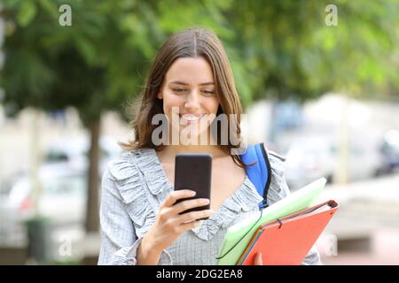 Vista frontale ritratto di uno studente felice che cammina controllando intelligente telefono in strada Foto Stock