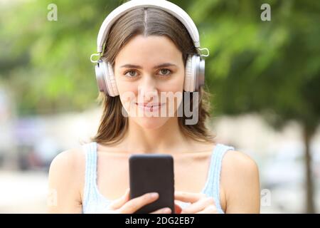 Vista frontale di una donna felice che indossa cuffie che tengono il cellulare telefono che ti guarda in strada Foto Stock