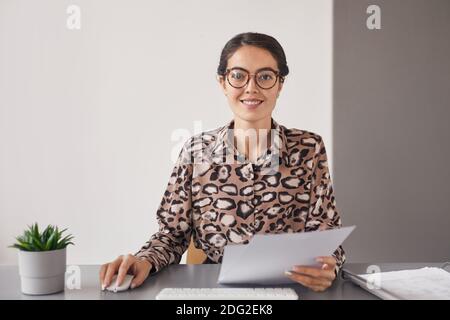 Ritratto minimo di giovane donna d'affari che indossa occhiali e sorridente alla fotocamera mentre si siede sul posto di lavoro tenendo un documento, spazio di copia Foto Stock
