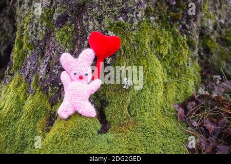 Coniglietto giocattolo lavorato a maglia con cuore decorativo rosso su muschio verde nella foresta. Foto Stock