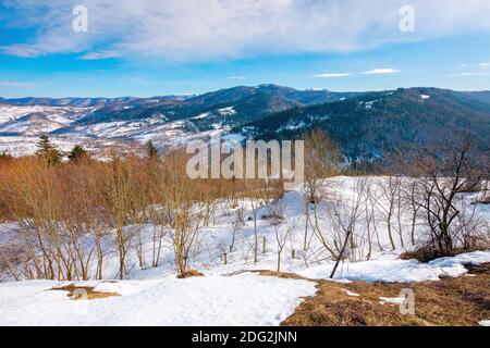 mountainous countryside on a sunny day. late winter scenery or beginning of spring. melting snow and leafless trees on the hills. village in the dista Foto Stock