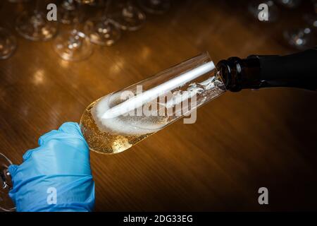 La mano blu del barman tiene un bicchiere di spumante. Lo champagne viene versato in un bicchiere. Festa durante la pandemia. Foto Stock