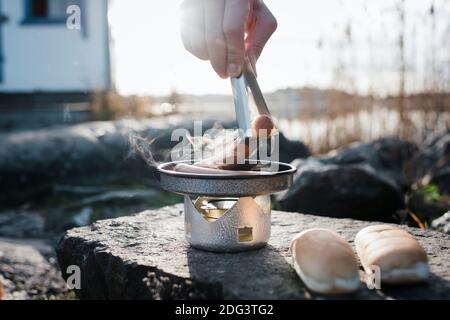 tenendolo a mano, si cuoce una salsiccia su un fornello da campeggio durante le escursioni Foto Stock