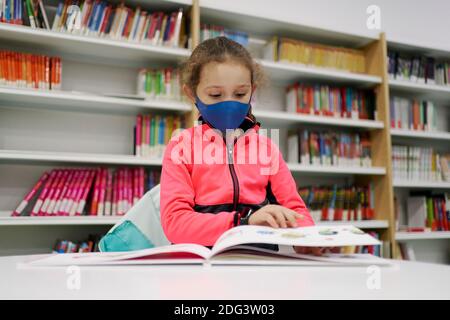 Bambina che indossa una maschera medica e che legge un libro in una biblioteca. Concetto di ritorno a scuola Foto Stock