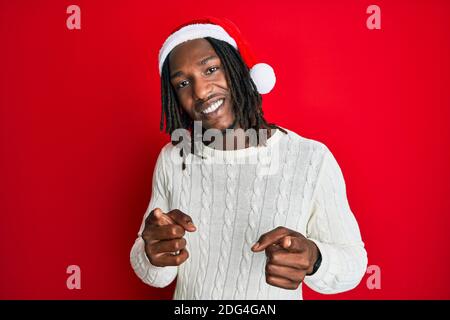 Uomo afro-americano con trecce che indossano cappello di natale puntando le dita alla macchina fotografica con faccia felice e divertente. Buona energia e vibrazioni. Foto Stock