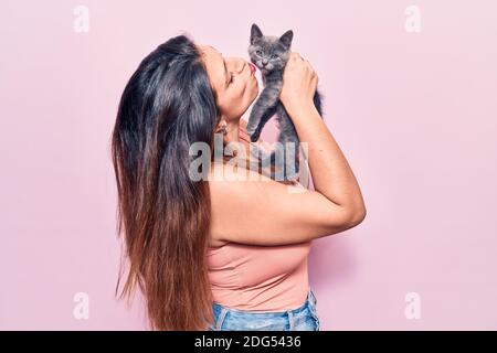 Giovane bella donna latina che puzzava felice. In piedi con sorriso sulla faccia che tiene e baciare adorabile gatto su isolato sfondo rosa Foto Stock