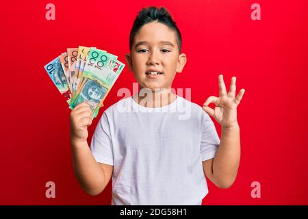 Piccolo ragazzo ispanico ragazzo che tiene dollari australiani facendo ok segno con le dita, sorridente amichevole gesturing eccellente simbolo Foto Stock