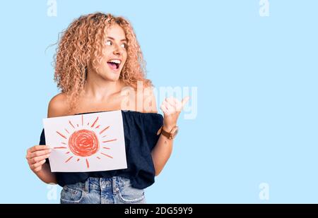 Giovane donna bionda con capelli ricci che tengono il sole disegnare puntando pollice fino al lato sorridente felice con bocca aperta Foto Stock