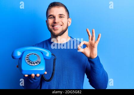 Giovane bell'uomo che tiene il telefono vintage facendo ok segno con le dita, sorridente amichevole gesturing simbolo eccellente Foto Stock