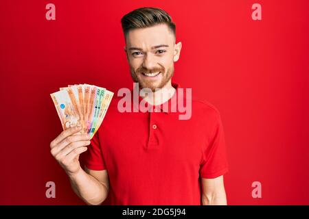 Giovane uomo redhead che tiene le banconote di peso filippino che osservano positive e. felice in piedi e sorridente con un sorriso sicuro che mostra i denti Foto Stock