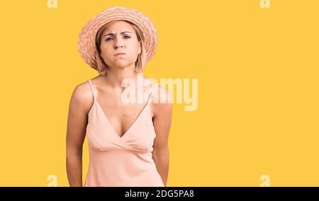 Giovane donna bionda con cappello estivo che soffia le guance con il viso divertente. Bocca gonfia con aria, espressione pazza. Foto Stock