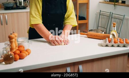 La donna più anziana chef impasta l'impasto sul tavolo in una cucina moderna. Panettiere anziano in pensione con bonete mescolando gli ingredienti con farina di grano setacciata impastando per cuocere la torta tradizionale e il pane. Foto Stock
