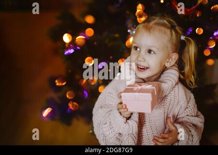 Allegra bambina di 4 anni, con una scatola regalo sullo sfondo di un festoso albero di Natale incandescente la vigilia di Capodanno Foto Stock