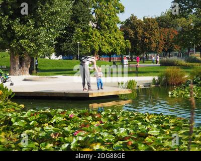 La mamma con la sua bimba sta guardando il lago verde con gigli d'acqua rosa e anatre al parco Bikás di Budapest, in Ungheria, in una giornata soleggiata di settembre Foto Stock