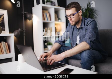 Uomo punteggiato in occhiali usando un computer portatile moderno per lavorare a distanza da casa. Concetto di freelance e tecnologia. Foto Stock