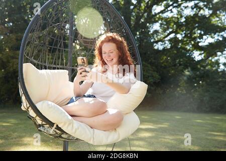 Felice ragazza di preteen che usa il telefono intelligente in sedia di swing dentro cortile soleggiato Foto Stock