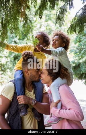 Coppia affettuosa che porta figlie sulle spalle sotto gli alberi del parco Foto Stock