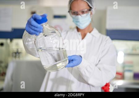 Donna scienziata in maschera facciale e guanto che esamina il liquido in laboratorio Foto Stock