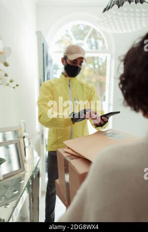 Donna che riceve consegna a casa da uomo in maschera con Smart telefono Foto Stock