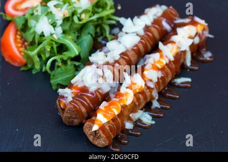 Cibo a base di spazzatura americana in una composizione semplice. Salsicce fritte croccanti o hot dog con salsa mayo e ketchup e cipolla tritata. Alcune verdure intorno lik Foto Stock