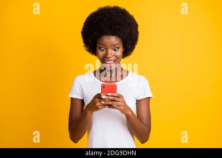 Ritratto fotografico di una donna afro-americana sbalordita che tiene il telefono in entrambe le mani indossare una t-shirt bianca isolata in giallo vivo colorato Foto Stock