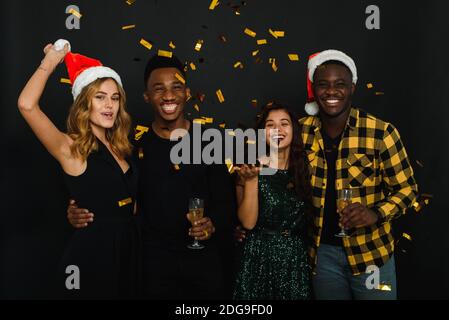 Quattro amici di diverse nazionalità gettano i confetti in cappelli e formiche babbo natale e bevono champagne su sfondo nero. Un'azienda di yo Foto Stock