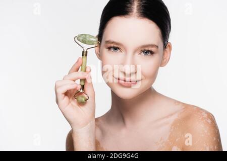 sorridente giovane bella donna con vitiligo che tiene rullo di giada sopra faccia isolata su bianco Foto Stock