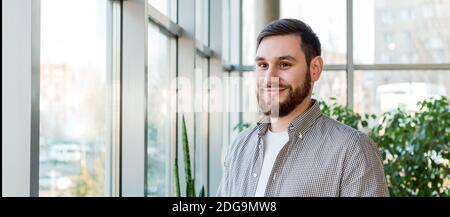 Sorridente uomo caucasico in piedi vicino a una finestra a lunghezza piena in un moderno ufficio con piante. Giovane bearded riuscito. Bell'uomo d'affari intelligente dentro Foto Stock