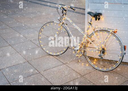 Luci di Natale decorate in bicicletta nella piazza della città. Bici bianca d'epoca con luci natalizie e fiocchi di neve che cadono. Umore di Natale Foto Stock