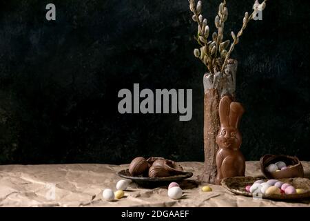 Umore di Pasqua ancora vita con rami di salice fiorito in vaso di ceramica, coniglio di cioccolato tradizionale, uova e dolci sul tavolo con carta da artigianato crumpled. Foto Stock