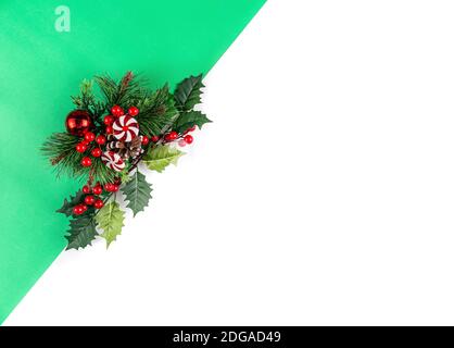 Sfondo verde e bianco. Rami realistici dell'albero di Natale con l'agrifoglio, le foglie, la palla rossa e il lollipop. Disposizione piatta. Foto Stock