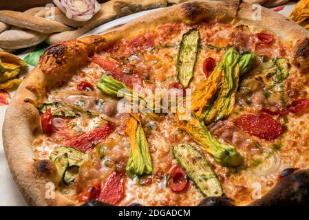 pizza con zucchine a fette, fiori di zucchine, pomodori e prosciutto, appetitosa pizza rotonda con vista dall'alto sul bordo croccante, primo piano Foto Stock