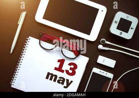 13 maggio. Giorno 13 del mese, data del calendario. Ambiente di lavoro in ufficio con notebook, notebook, forniture per ufficio e cancelleria sul retro marrone. Mese di primavera, giorno di t Foto Stock