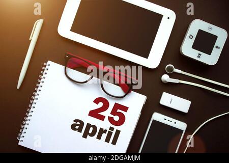 25 aprile. Giorno 25 del mese, data del calendario. Ambiente di lavoro in ufficio con notebook, notebook, forniture per ufficio e cancelleria sul retro marrone. Mese di primavera, giorno di Foto Stock