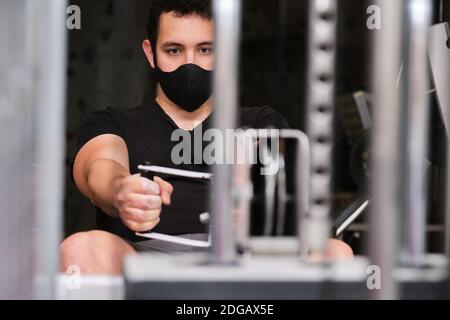 Fitness giovane uomo che indossa una maschera protettiva per il viso utilizzando la macchina da rowing in palestra. Nuovo esercizio normale. Foto Stock