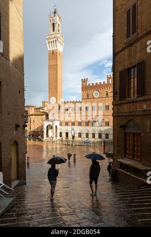 Viottolo verso la Torre del Mangia nel tempo con i turisti che tengono in primo piano gli ombrelloni, Siena, Toscana, Italia Foto Stock
