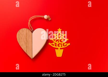 Medaglione di cuore di legno di due lati separati posto accanto ad un albero di Natale di carta giallo tagliato fuori, isolato su sfondo rosso luminoso con spazio di copia Foto Stock