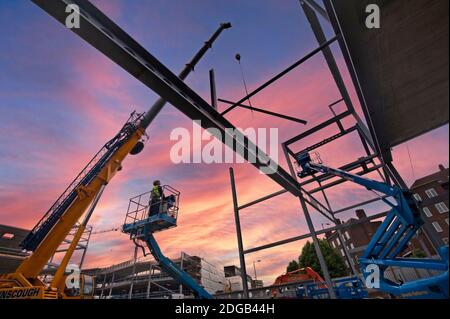 COSTRUIRE il Regno Unito BRITISH COSTRUZIONE DAWN GRU TRAVE CANTIERE LAVORATORI sorgere sopra cantiere edile, con cherry picker piattaforma di sollevamento operatore si prepara a controllare il paranco di una grande trave in metallo Foto Stock